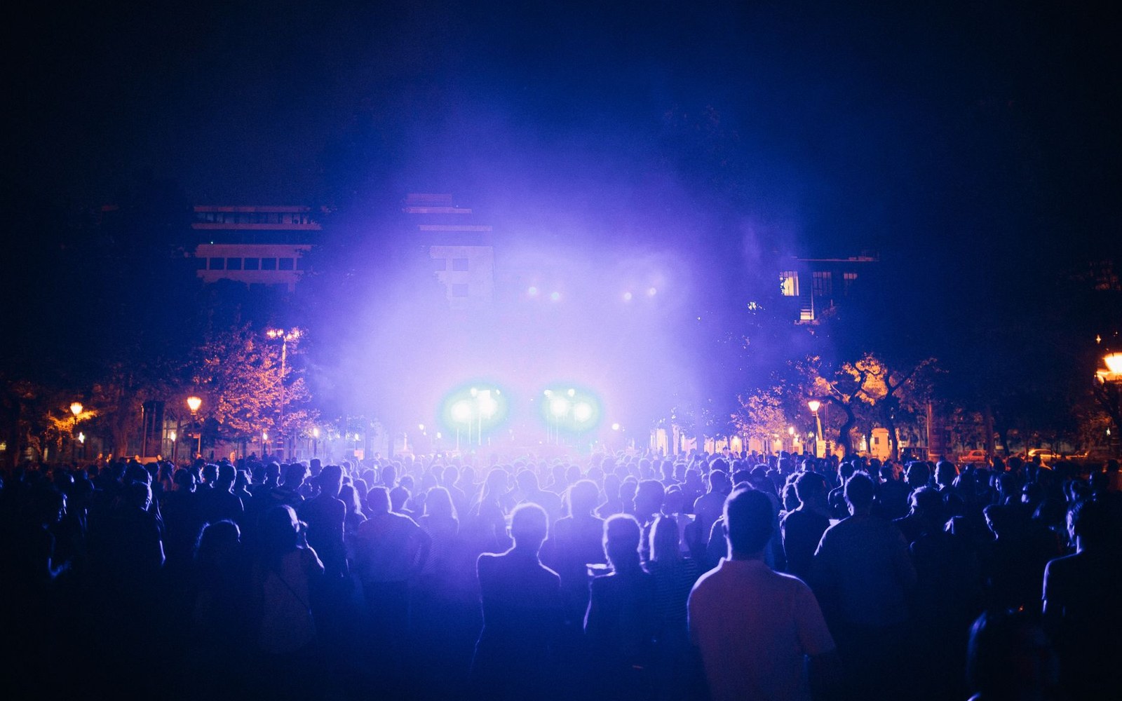 Festival crowd at dusk with a lit stage rig visible behind the performer silhouette, adults only