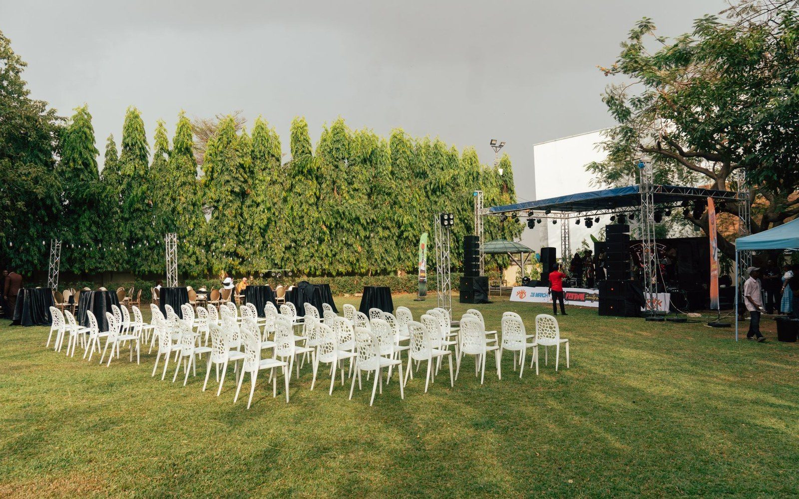 Rows of empty white chairs face an outdoor stage under a cloudy sky