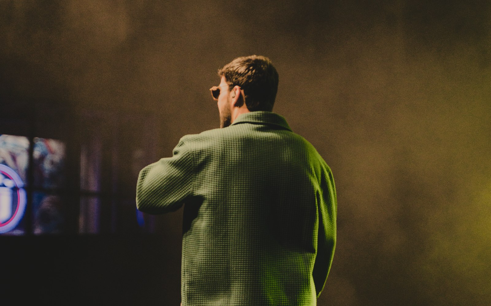 Man in green textured jacket seen from behind on a hazy, dimly lit stage