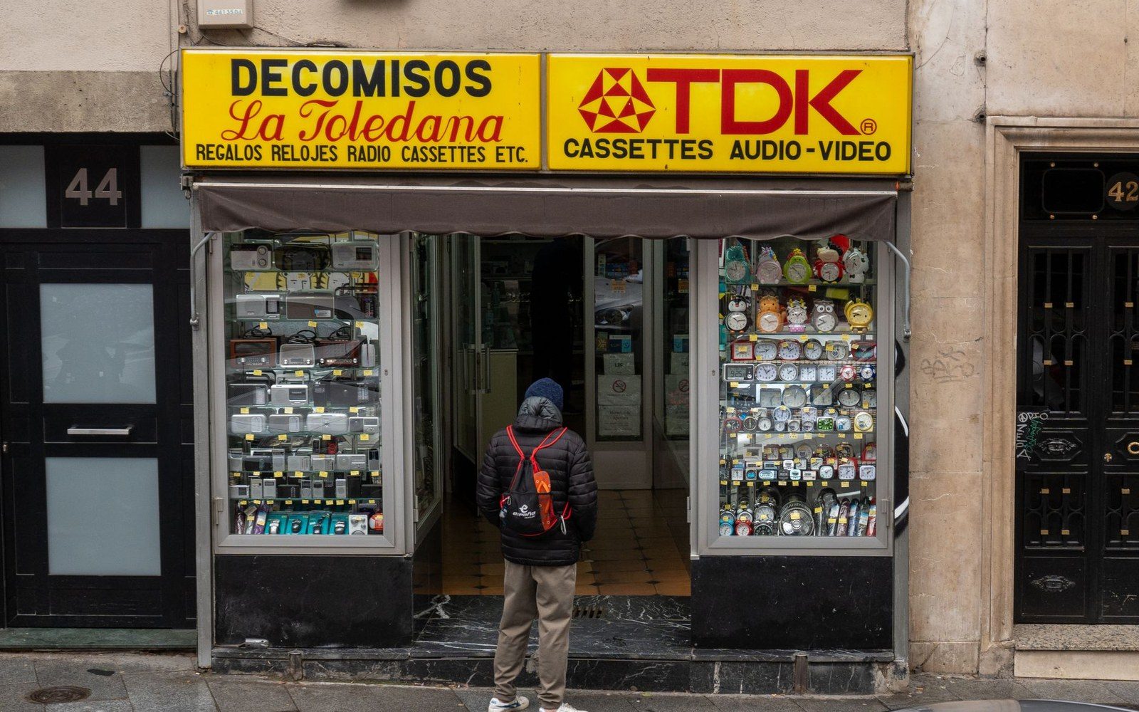 A pedestrian pauses outside a storefront advertising watches, radios, and TDK cassettes