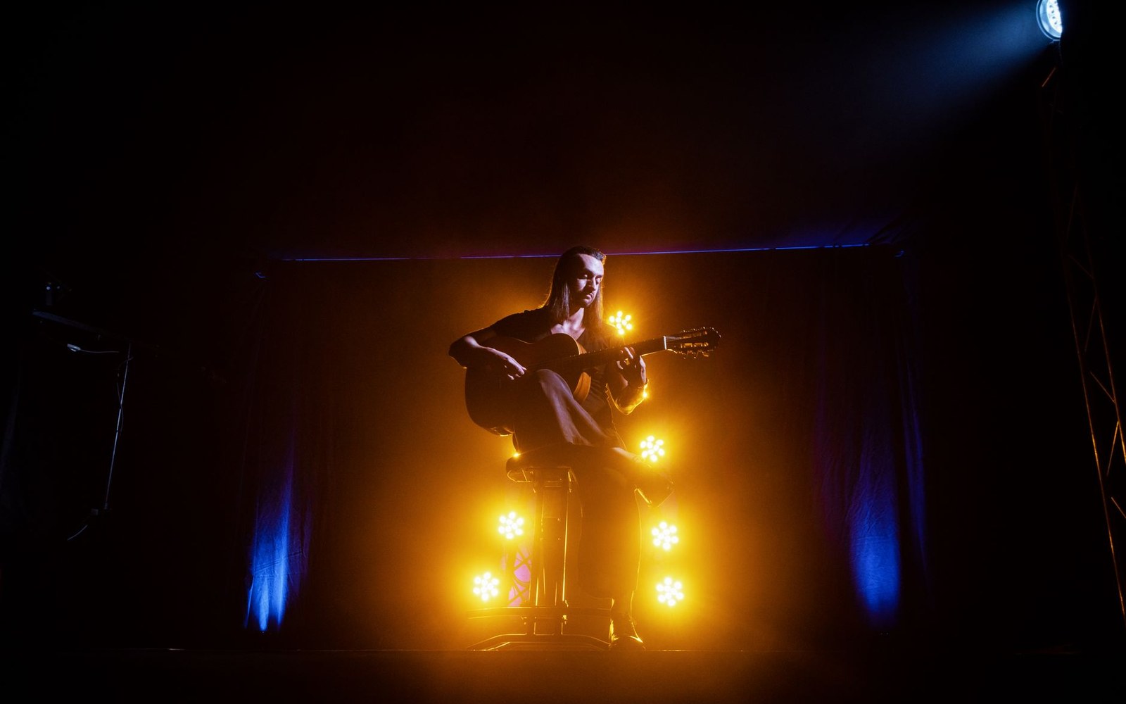 A guitarist sits on a stool amid glowing amber stage lights during a dark concert performance