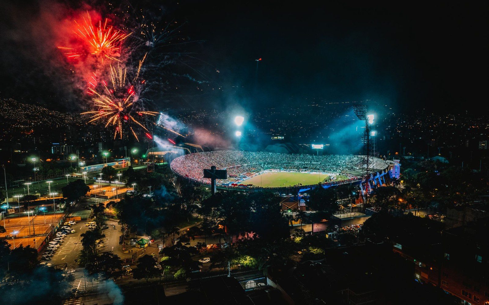 Fireworks light up the night sky over a packed illuminated stadium in a hillside city
