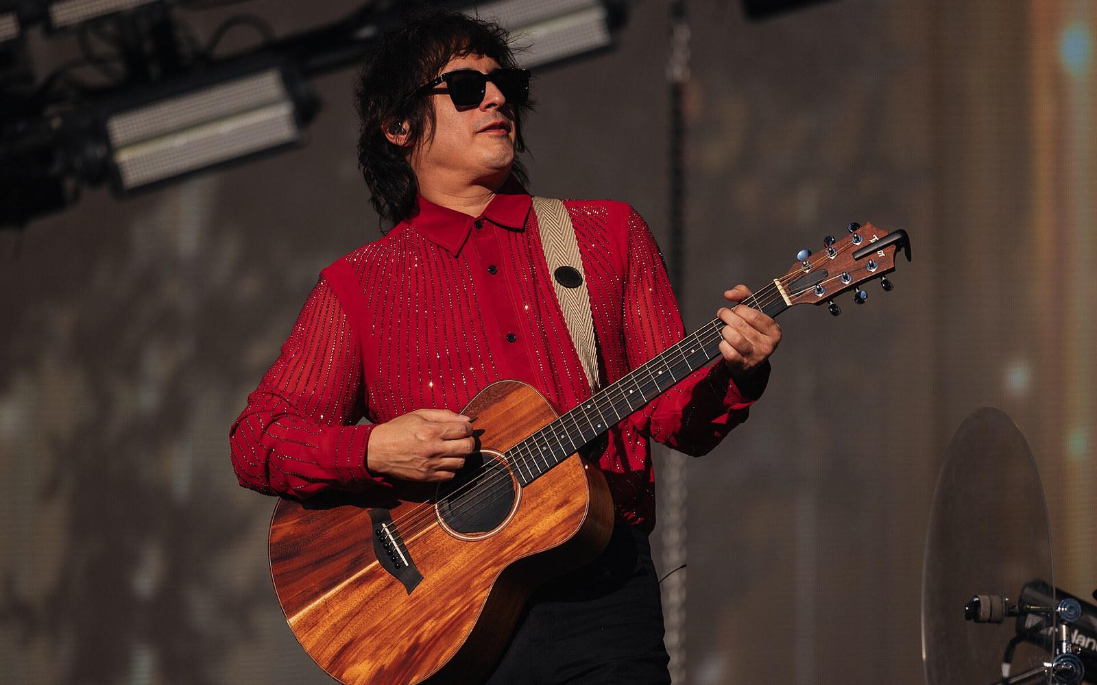 Musician in a sparkly red shirt and sunglasses plays acoustic guitar onstage