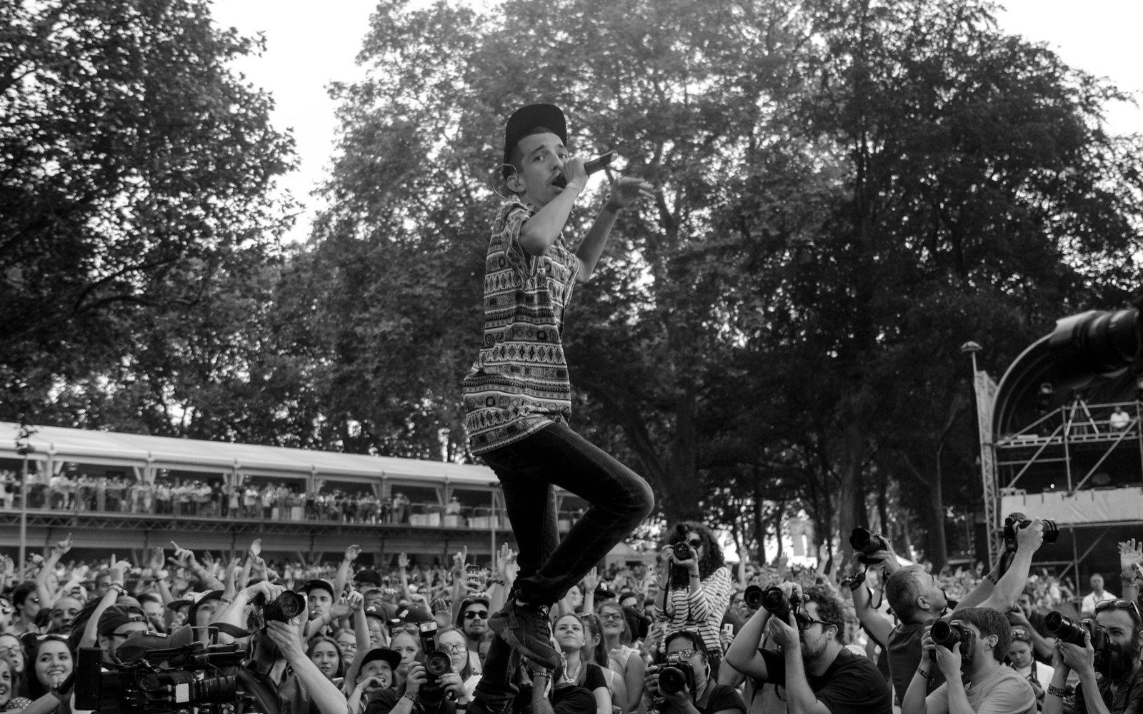 A performer sings into a microphone above a crowd of photographers and fans at an outdoor festival
